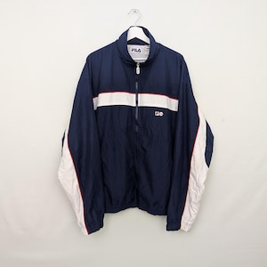 May include: Navy blue FILA track jacket with a white stripe across the chest and white accents on the sleeves. The jacket has a full zipper and the FILA logo on the chest. The jacket is hanging on a white hanger.