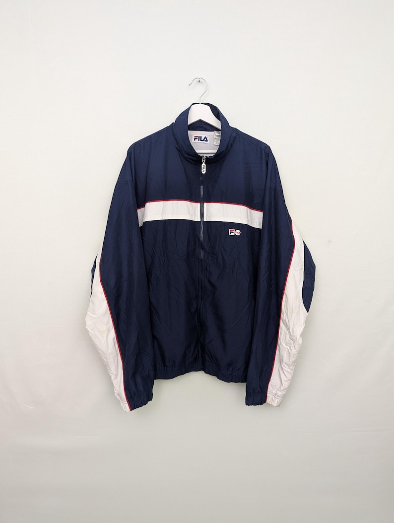 May include: Navy blue FILA track jacket with a white stripe across the chest and white accents on the sleeves. The jacket has a full zipper and the FILA logo on the chest. The jacket is hanging on a white hanger.