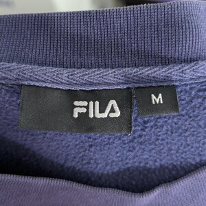 May include: Close-up of a purple FILA sweatshirt label. The label is black with the white FILA logo and the letter "M" on a separate tag. The sweatshirt has a ribbed neckline.