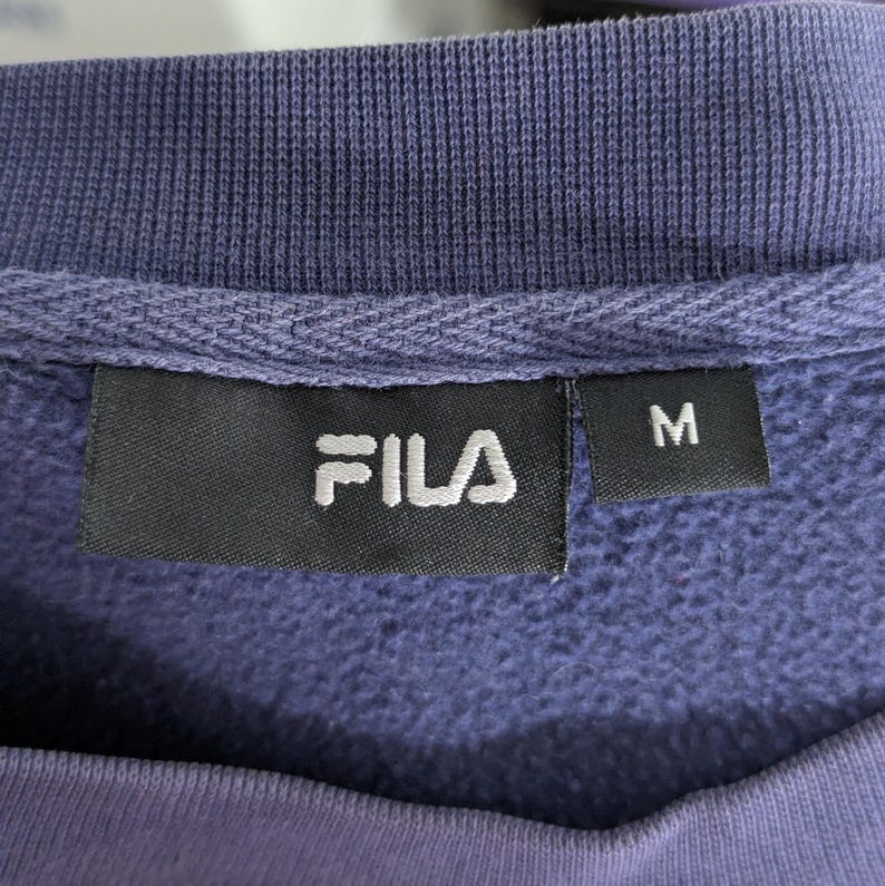 May include: Close-up of a purple FILA sweatshirt label. The label is black with the white FILA logo and the letter "M" on a separate tag. The sweatshirt has a ribbed neckline.