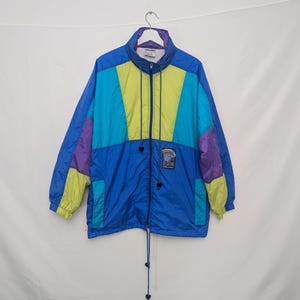 May include: A vintage windbreaker jacket in a vibrant colour block design. The jacket features a blue base with panels of turquoise, yellow, and purple. It has a high collar, a zip closure, and a small patch detail. The jacket is hanging against a white background.