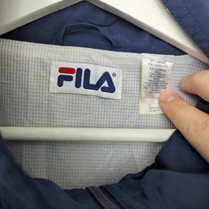 May include: Navy blue FILA jacket with a white and blue logo. The jacket is labeled XX-Large. The interior lining is light gray and white striped. The jacket is hanging on a white hanger.