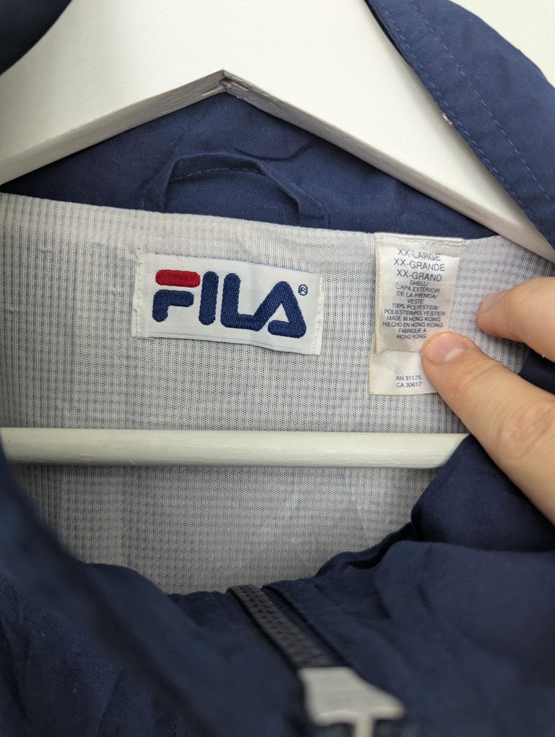 May include: Navy blue FILA jacket with a white and blue logo. The jacket is labeled XX-Large. The interior lining is light gray and white striped. The jacket is hanging on a white hanger.