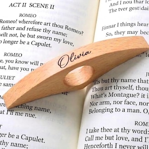 May include: A light-coloured wooden thumb page holder with a smooth, curved design and a central circular opening. The name Olivia is engraved in a dark, elegant script font on the top surface of the wood, resting on an open book page.