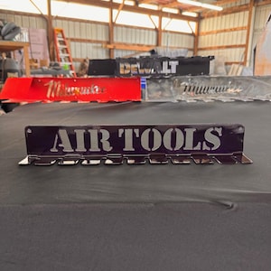 Puede incluir: Un soporte de herramientas de metal púrpura con las palabras "AIR TOOLS" en letras plateadas. El soporte tiene varias ranuras para herramientas y se muestra sobre una superficie negra. Otros soportes de herramientas son visibles en el fondo.