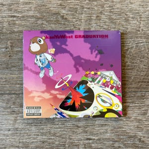 May include: A square music CD case featuring the album cover for Kanye West's Graduation. The artwork displays a brown bear character floating in a purple sky above a colorful, abstract landscape with a building and a Parental Advisory label.