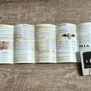 May include: A vintage cassette tape with a folded booklet featuring song lyrics and artwork. The cassette is off-white with a black label. The booklet has illustrations and text.