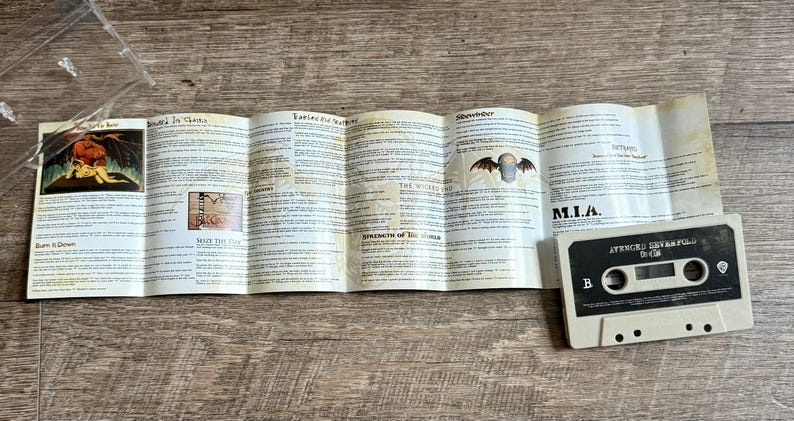 May include: A vintage cassette tape with a folded booklet featuring song lyrics and artwork. The cassette is off-white with a black label. The booklet has illustrations and text.