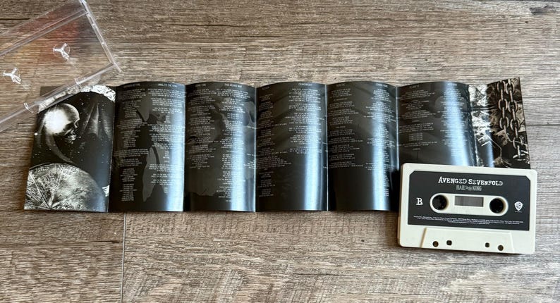 May include: A cassette tape labeled "Avenged Sevenfold Hail to the King" with a black and white design. The album insert is open, displaying lyrics and artwork. A clear plastic cassette case is partially visible.