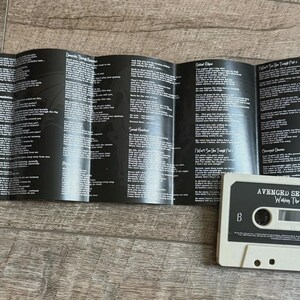 May include: A cassette tape and lyric sheet from the band Avenged Sevenfold. The cassette is off-white with black text, and the lyric sheet is black with white text. The cassette tape says "Avenged Sevenfold" and "Waking the Fallen".