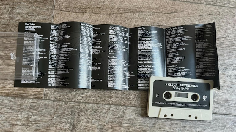 May include: A cassette tape and lyric sheet from the band Avenged Sevenfold. The cassette is off-white with black text, and the lyric sheet is black with white text. The cassette tape says "Avenged Sevenfold" and "Waking the Fallen".