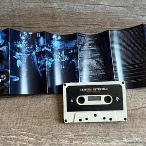 May include: A white cassette tape with the text "AVENGED SEVENFOLD NIGHTMARE" is centered on a wooden surface. The cassette is next to a folded booklet with blue and black artwork and lyrics.