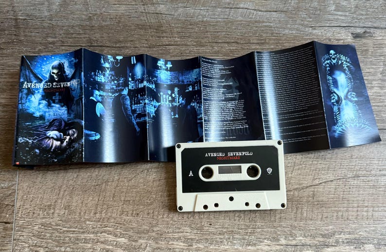 May include: A white cassette tape with the text "AVENGED SEVENFOLD NIGHTMARE" is centered on a wooden surface. The cassette is next to a folded booklet with blue and black artwork and lyrics.
