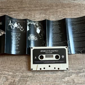 May include: A cassette tape and its unfolded insert are displayed on a wooden surface. The cassette is off-white with black text that reads "AVENGED SEVENFOLD HAIL TO THE KING". The insert is black with white text and graphics.