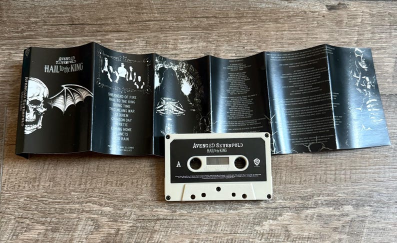 May include: A cassette tape and its unfolded insert are displayed on a wooden surface. The cassette is off-white with black text that reads "AVENGED SEVENFOLD HAIL TO THE KING". The insert is black with white text and graphics.
