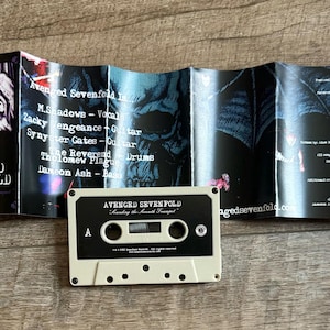 May include: An Avenged Sevenfold cassette tape and album art are displayed on a wooden surface. The cassette is off-white with black text. The album art features band member names and song lyrics.