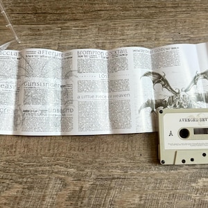 May include: A cassette tape labeled "AVENGED SEVENFOLD" sits on a wooden surface next to a folded paper with song lyrics and a winged skull graphic. The tape is light gray with black details.