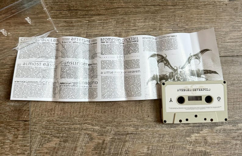 May include: A cassette tape labeled "AVENGED SEVENFOLD" sits on a wooden surface next to a folded paper with song lyrics and a winged skull graphic. The tape is light gray with black details.