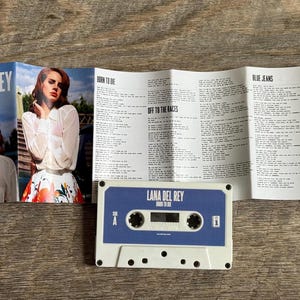 May include: A cassette tape and album art for Lana Del Rey's "Born to Die" album. The cassette is blue and white, with the album title printed on it. The album art features a photo of Lana Del Rey.