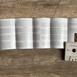 May include: A light gray cassette tape labeled "Lana Del Rey Blue Banisters" with a track list insert. The cassette is on a wooden surface, with the clear plastic case partially visible. The insert displays song lyrics.