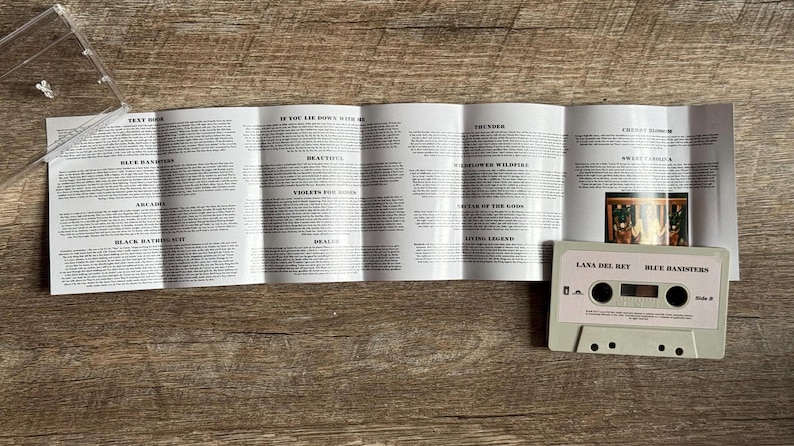 May include: A light gray cassette tape labeled "Lana Del Rey Blue Banisters" with a track list insert. The cassette is on a wooden surface, with the clear plastic case partially visible. The insert displays song lyrics.