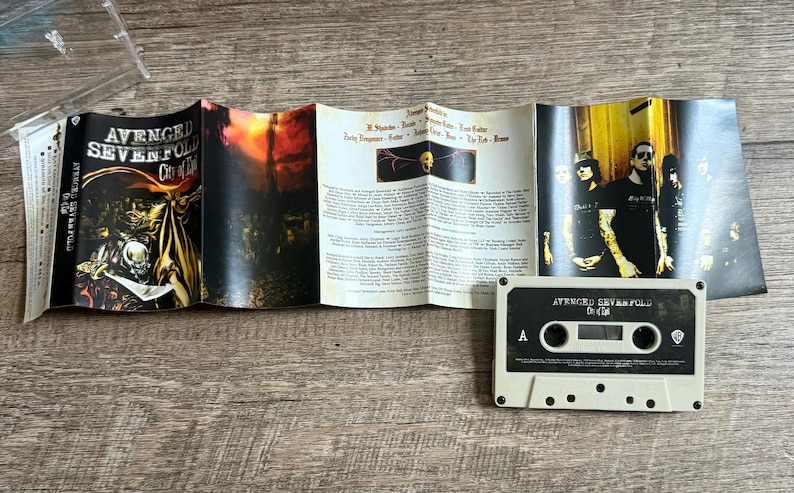 May include: A cassette tape and its accompanying booklet are displayed on a wooden surface. The cassette is labeled "Avenged Sevenfold City of Evil." The booklet features band photos and lyrics.