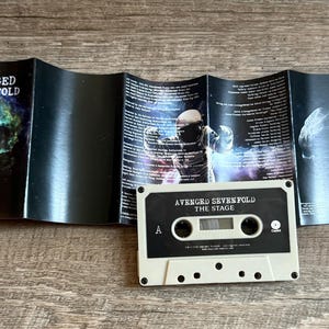 May include: A vintage cassette tape and its accompanying booklet are displayed on a wooden surface. The cassette is off-white with black accents and the text "AVENGED SEVENFOLD THE STAGE". The booklet features album art and lyrics.