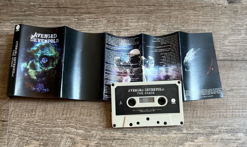 May include: A vintage cassette tape and its accompanying booklet are displayed on a wooden surface. The cassette is off-white with black accents and the text "AVENGED SEVENFOLD THE STAGE". The booklet features album art and lyrics.