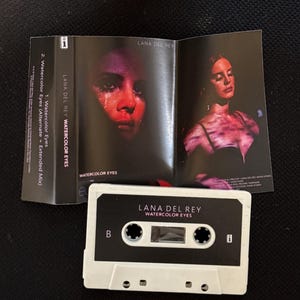 May include: A white cassette tape labeled Lana Del Rey Watercolor Eyes with a B side indicator. The accompanying paper insert features two photographs of a person with red lighting, the text Lana Del Rey Watercolor Eyes, and track listings.