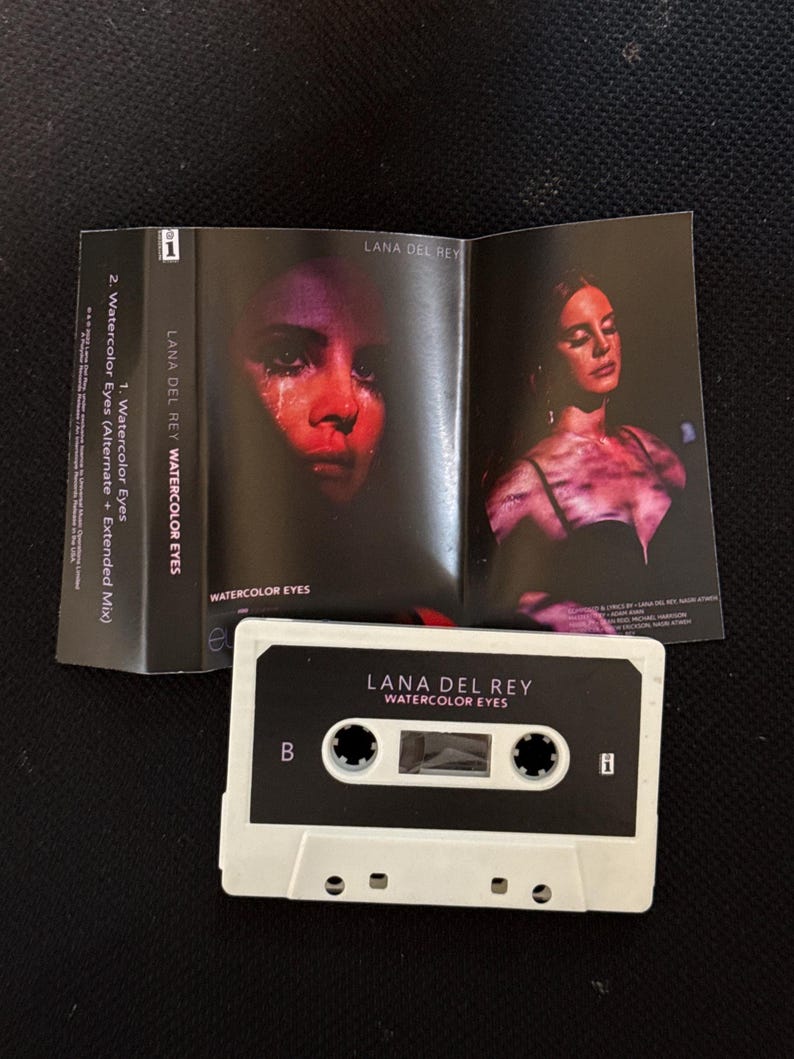 May include: A white cassette tape labeled Lana Del Rey Watercolor Eyes with a B side indicator. The accompanying paper insert features two photographs of a person with red lighting, the text Lana Del Rey Watercolor Eyes, and track listings.