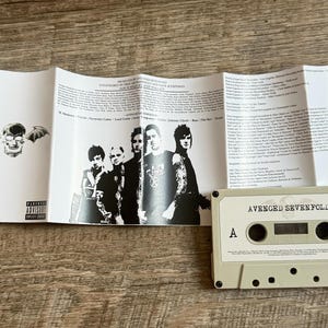 May include: A cassette tape and album insert featuring the band Avenged Sevenfold. The cassette is light beige with black details. The insert displays the band members, lyrics, and album art, including a skull with wings.