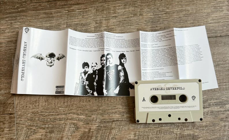 May include: A cassette tape and album insert featuring the band Avenged Sevenfold. The cassette is light beige with black details. The insert displays the band members, lyrics, and album art, including a skull with wings.