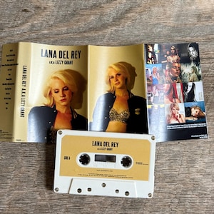 May include: A vintage cassette tape and album art for Lana Del Rey's album, "Lana Del Rey A.K.A. Lizzy Grant." The cassette is white with a tan label. The album art features photos of the artist.