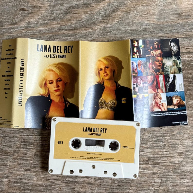 May include: A vintage cassette tape and album art for Lana Del Rey's album, "Lana Del Rey A.K.A. Lizzy Grant." The cassette is white with a tan label. The album art features photos of the artist.