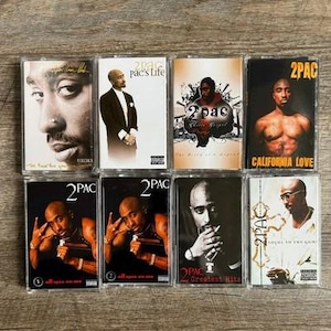 May include: A collection of eight 2Pac cassette tapes arranged in two rows. The covers feature various portraits of 2Pac, including titles like Pac's Life, Ghetto Gospel, California Love, All Eyez On Me, Greatest Hits, and Loyal To The Game with explicit content labels.