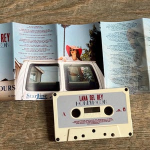 May include: A vintage cassette tape and album booklet for Lana Del Rey's "Honeymoon." The cassette is off-white with black details. The booklet features album art with a woman in a car and lyrics.