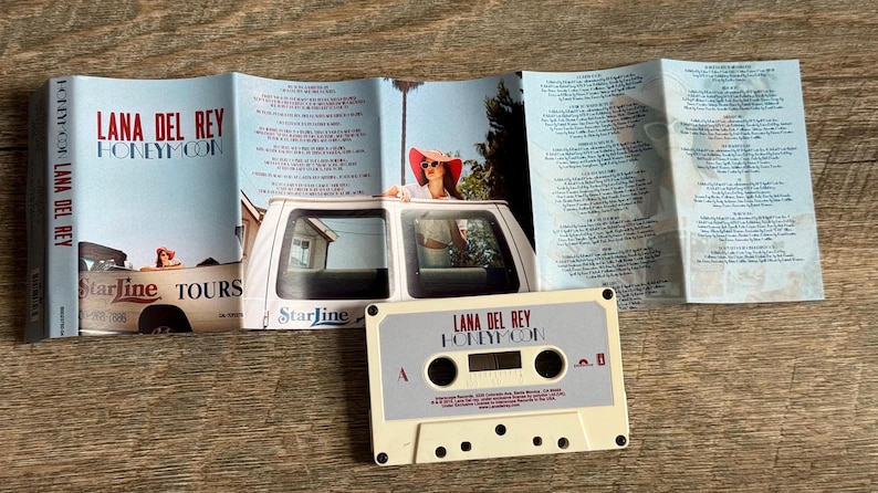 May include: A vintage cassette tape and album booklet for Lana Del Rey's "Honeymoon." The cassette is off-white with black details. The booklet features album art with a woman in a car and lyrics.