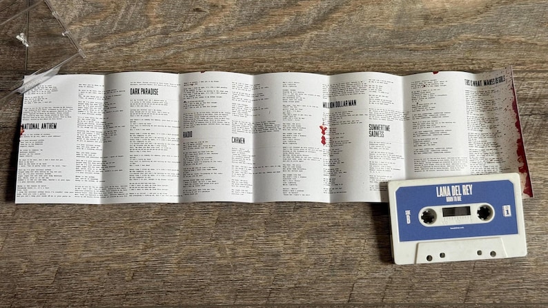 May include: A white cassette tape with a blue label featuring the text "LANA DEL REY BORN TO DIE" and a tracklist insert. The insert displays song lyrics. The cassette tape and insert are on a wooden surface.