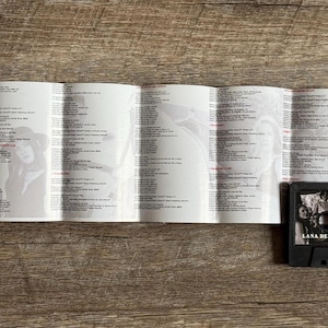 May include: A black cassette tape with the album title "Chemtrails Over the Country Club" and the artist name "Lana Del Rey" is displayed next to a folded booklet with lyrics and images. The items are on a wooden surface.