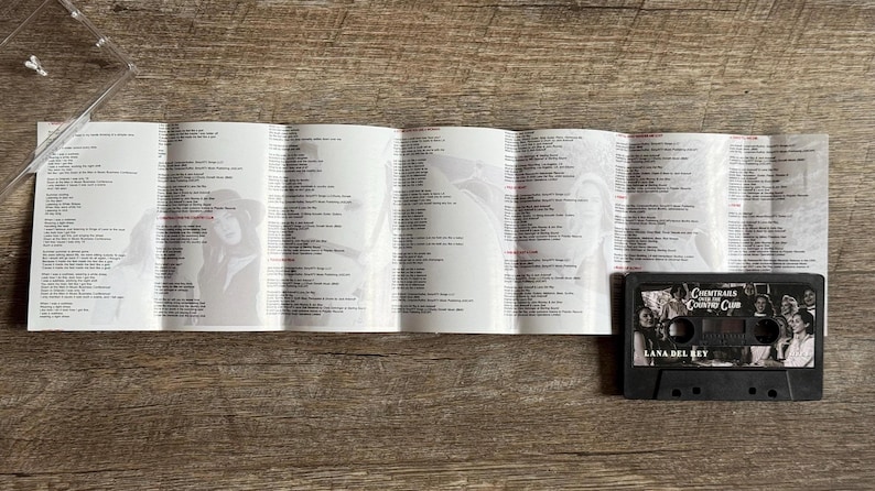 May include: A black cassette tape with the album title "Chemtrails Over the Country Club" and the artist name "Lana Del Rey" is displayed next to a folded booklet with lyrics and images. The items are on a wooden surface.