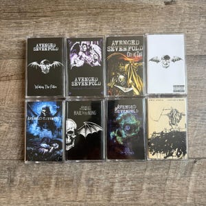 May include: A collection of eight cassette tapes featuring album art from the band Avenged Sevenfold. The tapes showcase various album covers with skulls, angels, and other gothic imagery. The album titles are visible on the cassette labels.