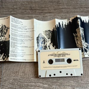 May include: A vintage cassette tape and its accompanying booklet are displayed on a wooden surface. The cassette is light beige with the text "AVENGED SEVENFOLD LIFE IS BUT A DREAM..." The booklet features black and white illustrations and song lyrics.