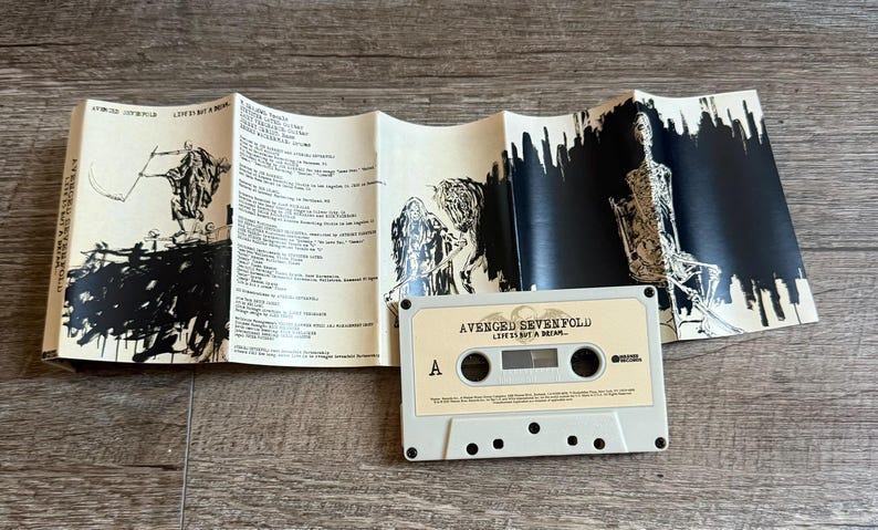 May include: A vintage cassette tape and its accompanying booklet are displayed on a wooden surface. The cassette is light beige with the text "AVENGED SEVENFOLD LIFE IS BUT A DREAM..." The booklet features black and white illustrations and song lyrics.