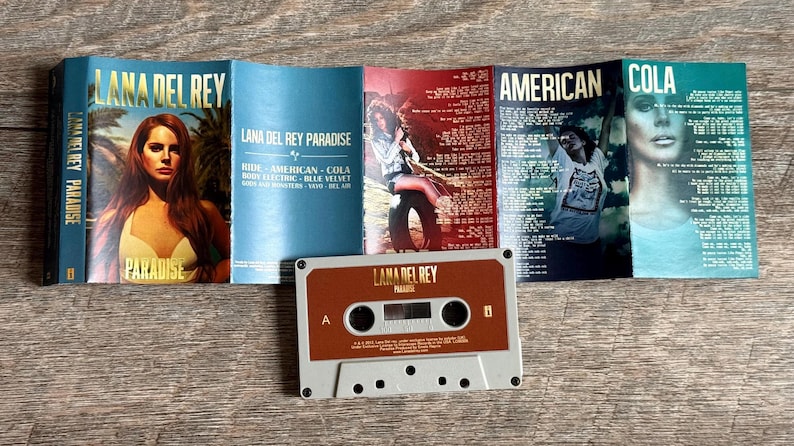 May include: A cassette tape and album art for Lana Del Rey's "Paradise" album. The cassette is brown and gray. The album art features the album title and a portrait of Lana Del Rey.