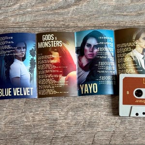 May include: A cassette tape and album art from Lana Del Rey's Paradise album. The cassette is brown and white. The album art features images of Lana Del Rey with song titles including "Body Electric", "Blue Velvet", "Gods & Monsters", and "Yayo".