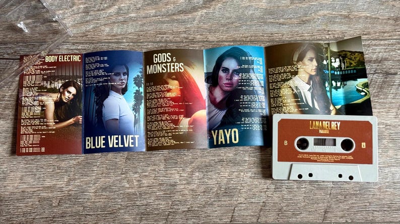 May include: A cassette tape and album art from Lana Del Rey's Paradise album. The cassette is brown and white. The album art features images of Lana Del Rey with song titles including "Body Electric", "Blue Velvet", "Gods & Monsters", and "Yayo".
