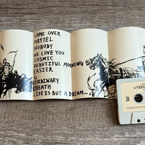 May include: A cassette tape with the text "AVENGED SEVENFOLD" and "LIFE IS BUT A DREAM" printed on it. The cassette is light gray. A folded insert with black illustrations and song titles is next to the cassette.