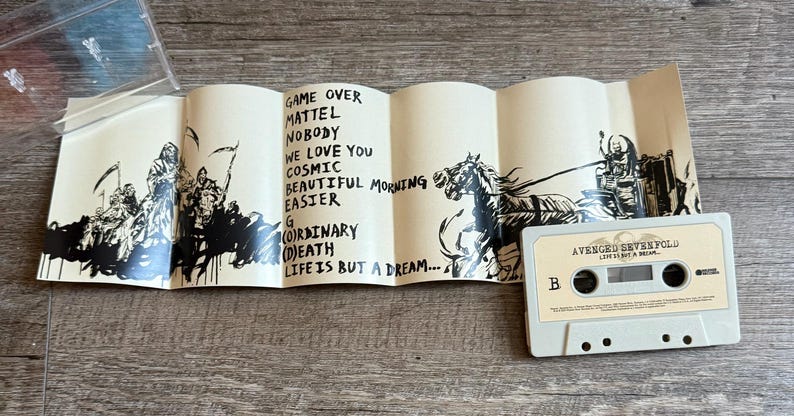 May include: A cassette tape with the text "AVENGED SEVENFOLD" and "LIFE IS BUT A DREAM" printed on it. The cassette is light gray. A folded insert with black illustrations and song titles is next to the cassette.