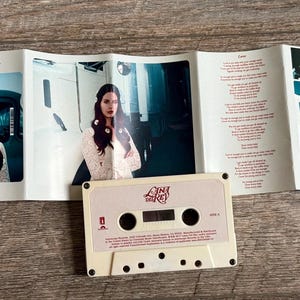 May include: A cassette tape and its accompanying booklet are displayed on a wooden surface. The cassette is off-white with the text "Lana Del Rey" and "Lust for Life." The booklet features photos of a woman and song lyrics.