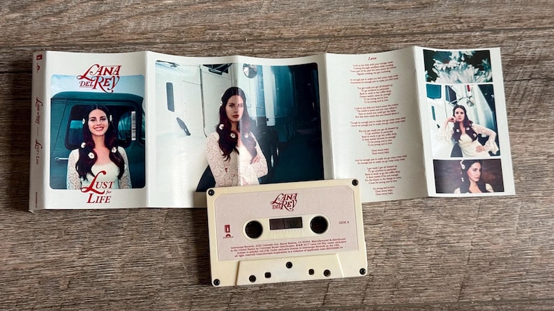 May include: A cassette tape and its accompanying booklet are displayed on a wooden surface. The cassette is off-white with the text "Lana Del Rey" and "Lust for Life." The booklet features photos of a woman and song lyrics.